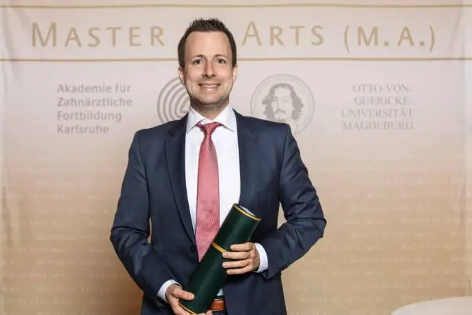 Ein Absolvent im Anzug hält eine grüne Urkundenrolle vor einer akademischen Logowand