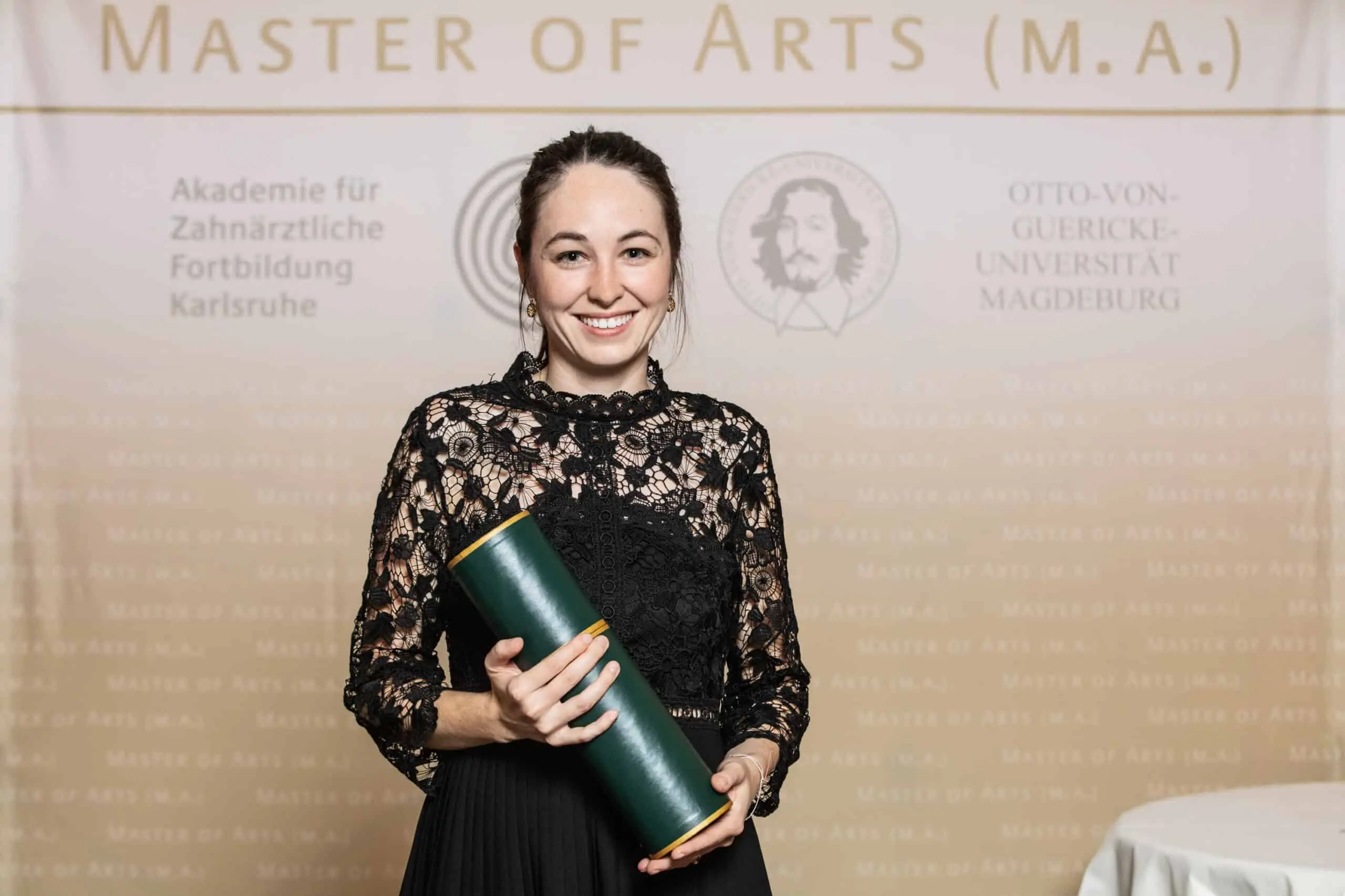 Eine lächelnde Absolventin in schwarzem Spitzenkleid hält eine grüne Zeugnisrolle vor einer Logowand mit dem Schriftzug Master of Arts.