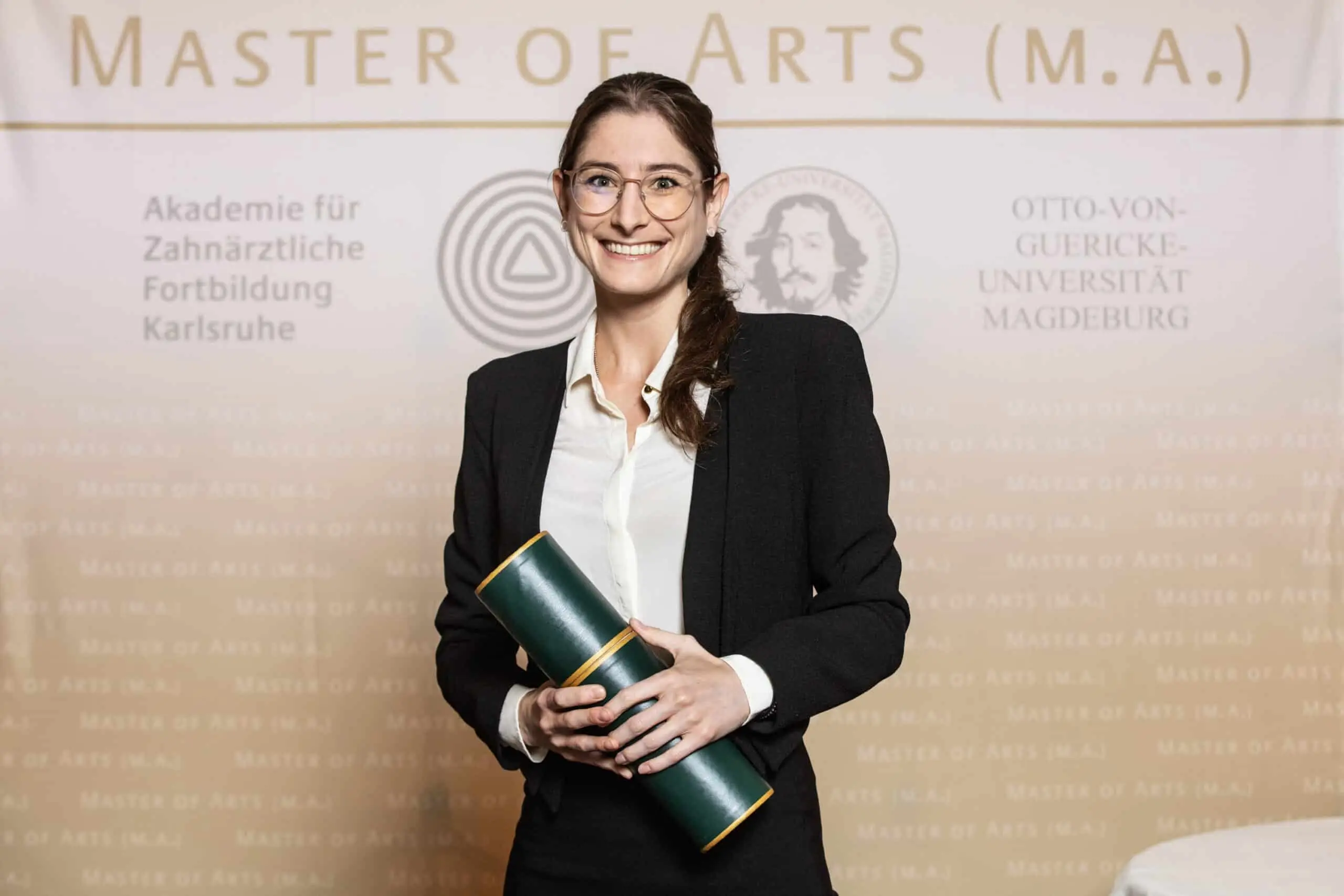 Eine lächelnde Absolventin in Business-Kleidung hält eine grüne Urkundenrolle vor einer hellen Fotowand mit akademischen Schriftzügen
