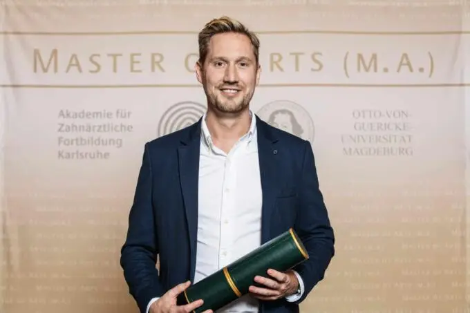 Ein lächelnder Mann im Business-Outfit hält eine grüne Zeugnisrolle vor einer hellen Wand mit akademischem Schriftzug