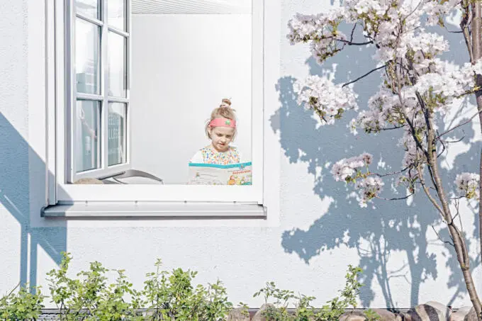 Kind liest Buch über Zahnärzte am offenen Fenster einer Praxis, Außenansicht mit blühendem Baum und Schattenwurf auf weißer Fassade