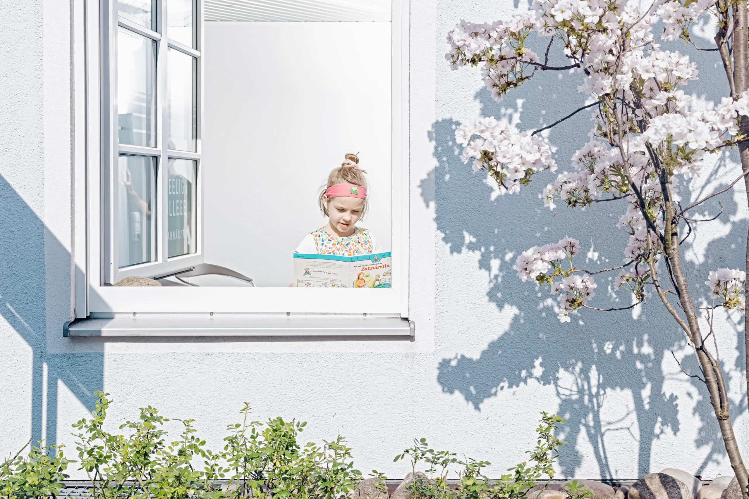 Kind liest Buch über Zahnärzte am offenen Fenster einer Praxis, Außenansicht mit blühendem Baum und Schattenwurf auf weißer Fassade