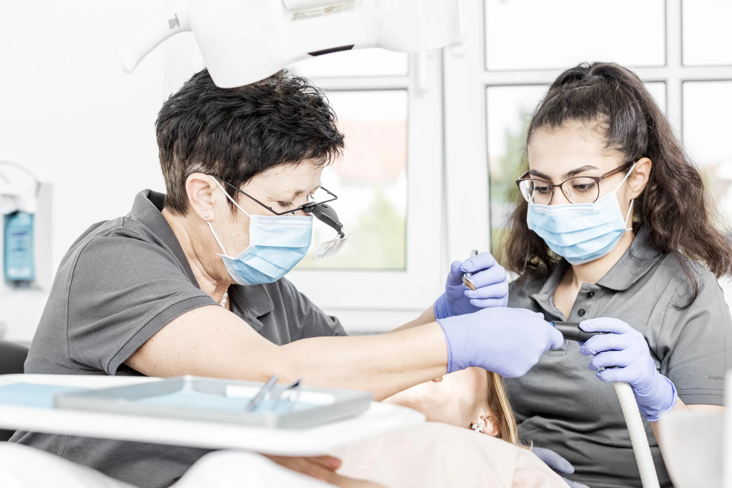 Zahnärztin mit Lupenbrille und Assistentin bei der Behandlung eines Patienten, beide mit Mundschutz und Handschuhen