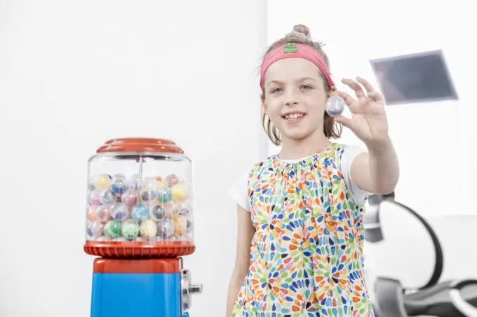 Lächelndes Mädchen zeigt Spielzeugkapsel neben einem Kaugummiautomaten in einer Zahnarztpraxis