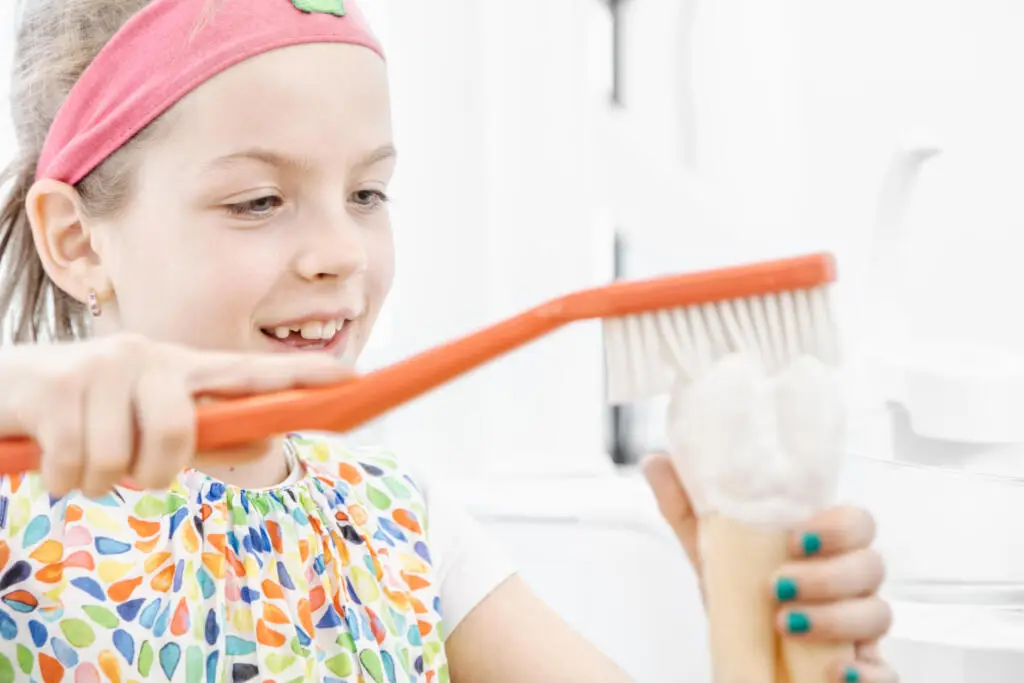 Mädchen mit rosa Stirnband übt Zähneputzen an einem Zahnmodell mit einer großen orangen Zahnbürste in einer hellen Praxisumgebung