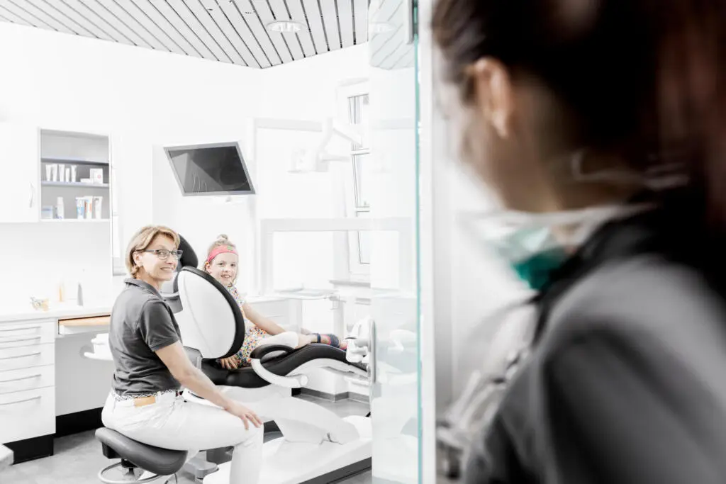 Blick durch eine Glastür auf eine lächelnde Zahnärztin und ein Kind im Behandlungsstuhl einer hellen Praxis
