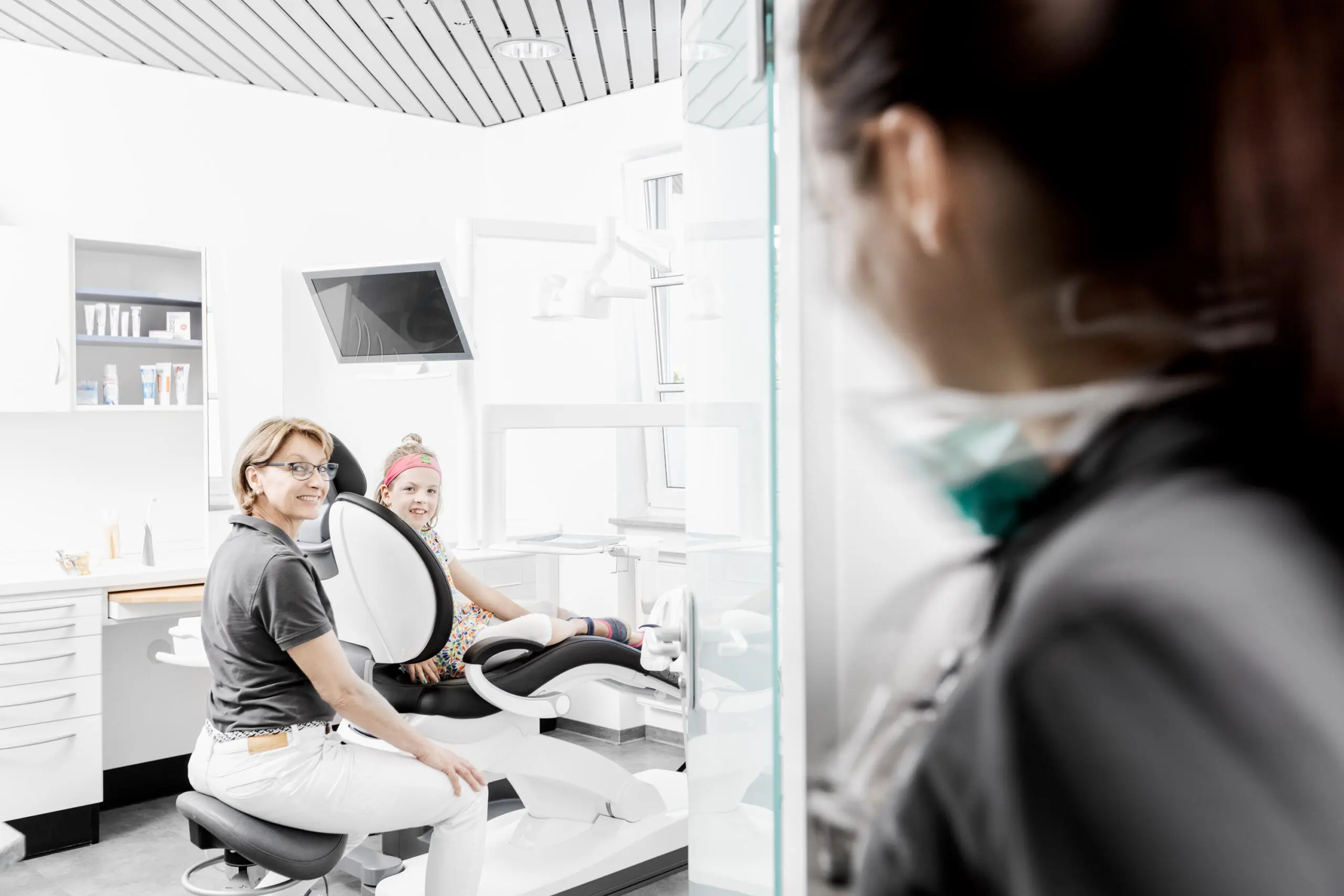Blick durch eine Glastür auf eine lächelnde Zahnärztin und ein Kind im Behandlungsstuhl einer hellen Praxis