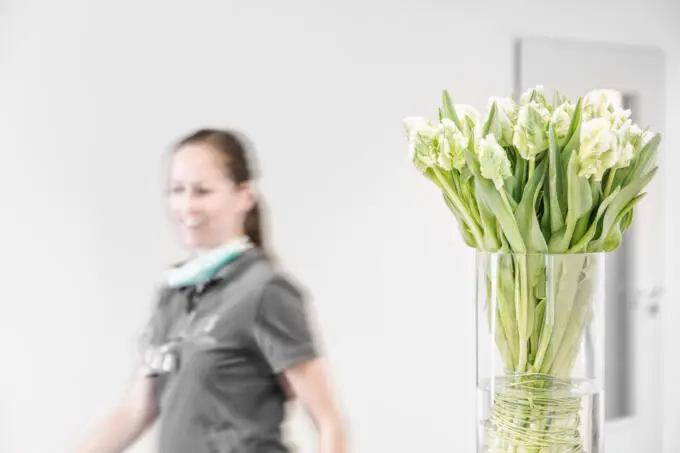 Weiße Tulpen in einer Glasvase im Fokus, im unscharfen Hintergrund eine zahnmedizinische Mitarbeiterin in grauer Arbeitskleidung