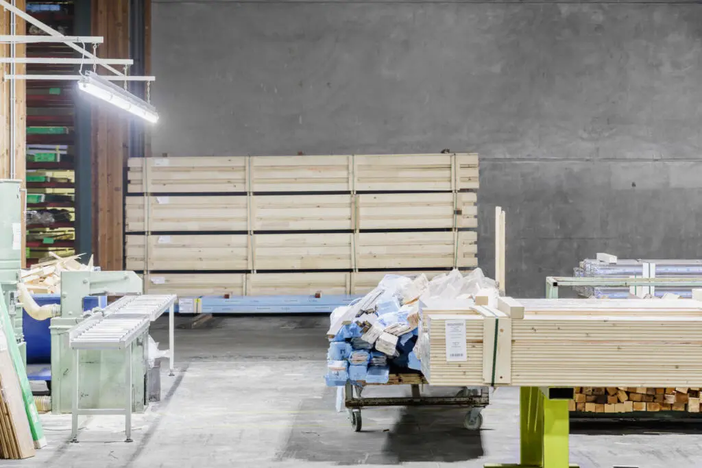 Innenansicht einer Lagerhalle mit gestapeltem Bauholz, einer grünen Bearbeitungsmaschine und Transportwagen vor einer grauen Betonwand.