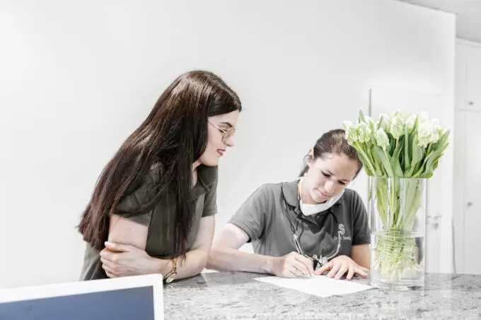 Zwei Mitarbeiterinnen in Arbeitskleidung am Empfangstresen einer Zahnarztpraxis beim Ausfüllen von Unterlagen mit weißen Tulpen im Vordergrund
