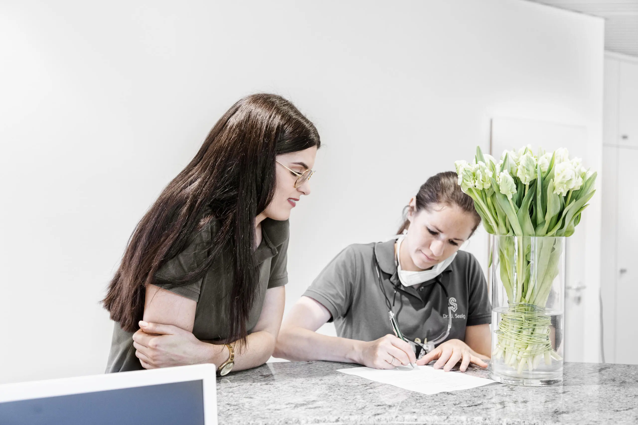 Zwei Mitarbeiterinnen in Arbeitskleidung am Empfangstresen einer Zahnarztpraxis beim Ausfüllen von Unterlagen mit weißen Tulpen im Vordergrund