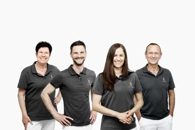 Vier Zahnärzte in einheitlichen grauen Poloshirts und weißen Hosen stehen lächelnd nebeneinander vor weißem Hintergrund