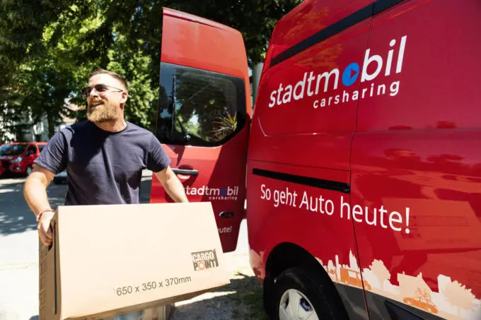 Mann trägt großen Karton neben rotem Carsharing-Transporter mit geöffneten Hecktüren bei Sonnenschein