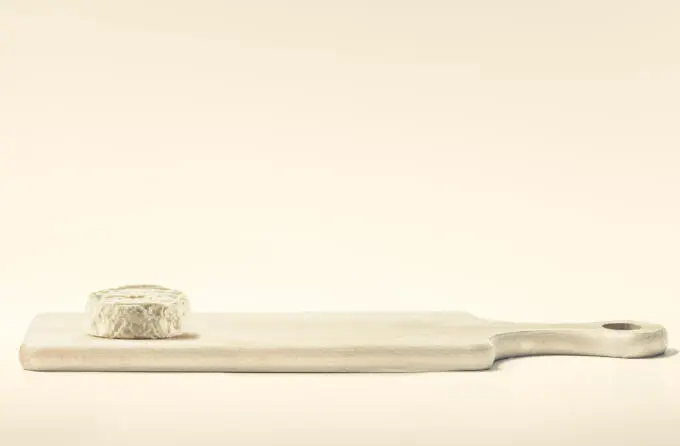 Ein runder Weichkäse liegt auf der linken Seite eines hellen Holzschneidebretts vor einem einfarbigen beigen Hintergrund.