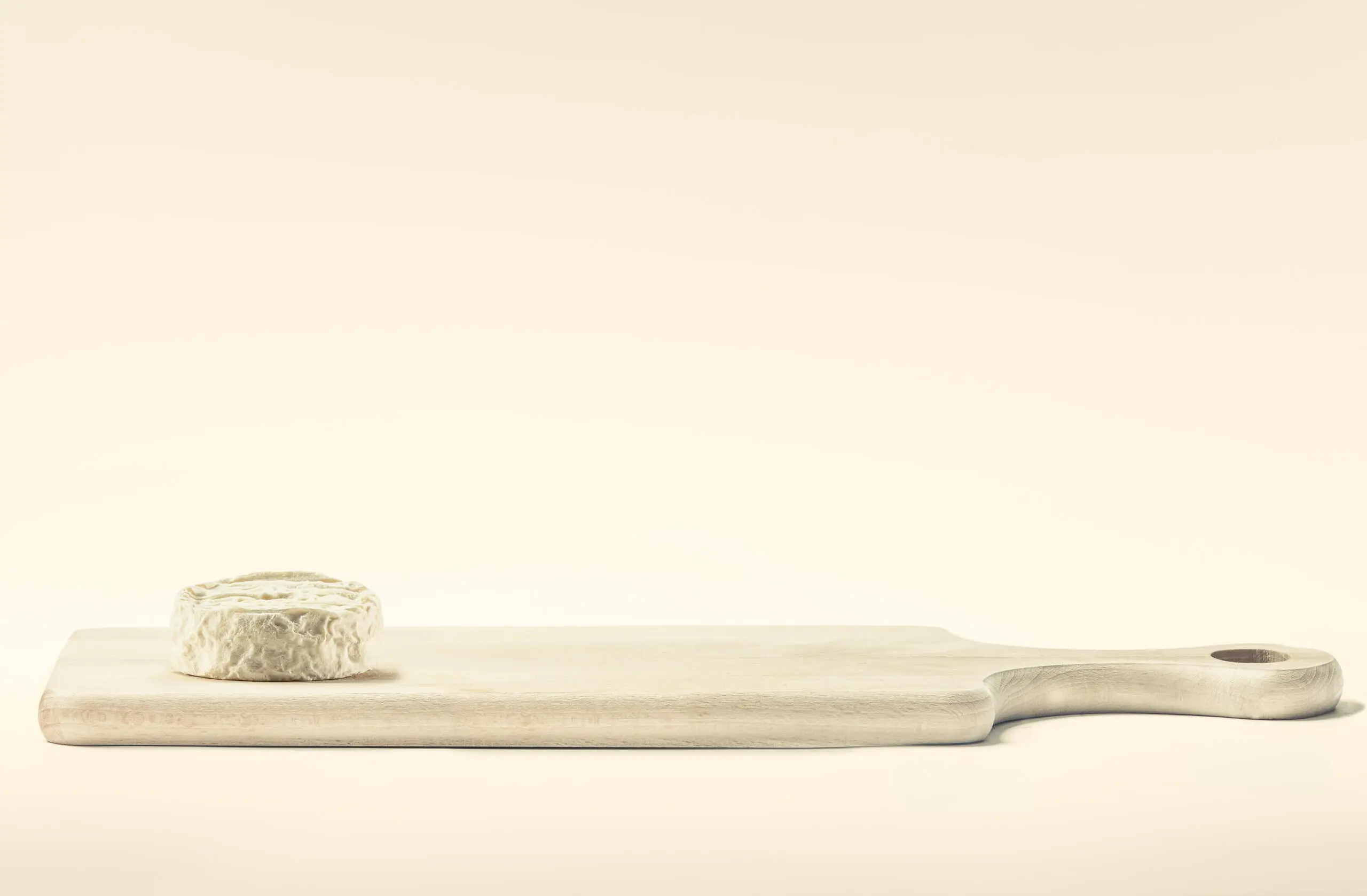 Ein runder Weichkäse liegt auf der linken Seite eines hellen Holzschneidebretts vor einem einfarbigen beigen Hintergrund.
