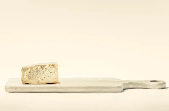Ein Stück cremiger Weichkäse liegt auf einem hellen Holzschneidebrett vor einem neutralen, cremefarbenen Hintergrund.
