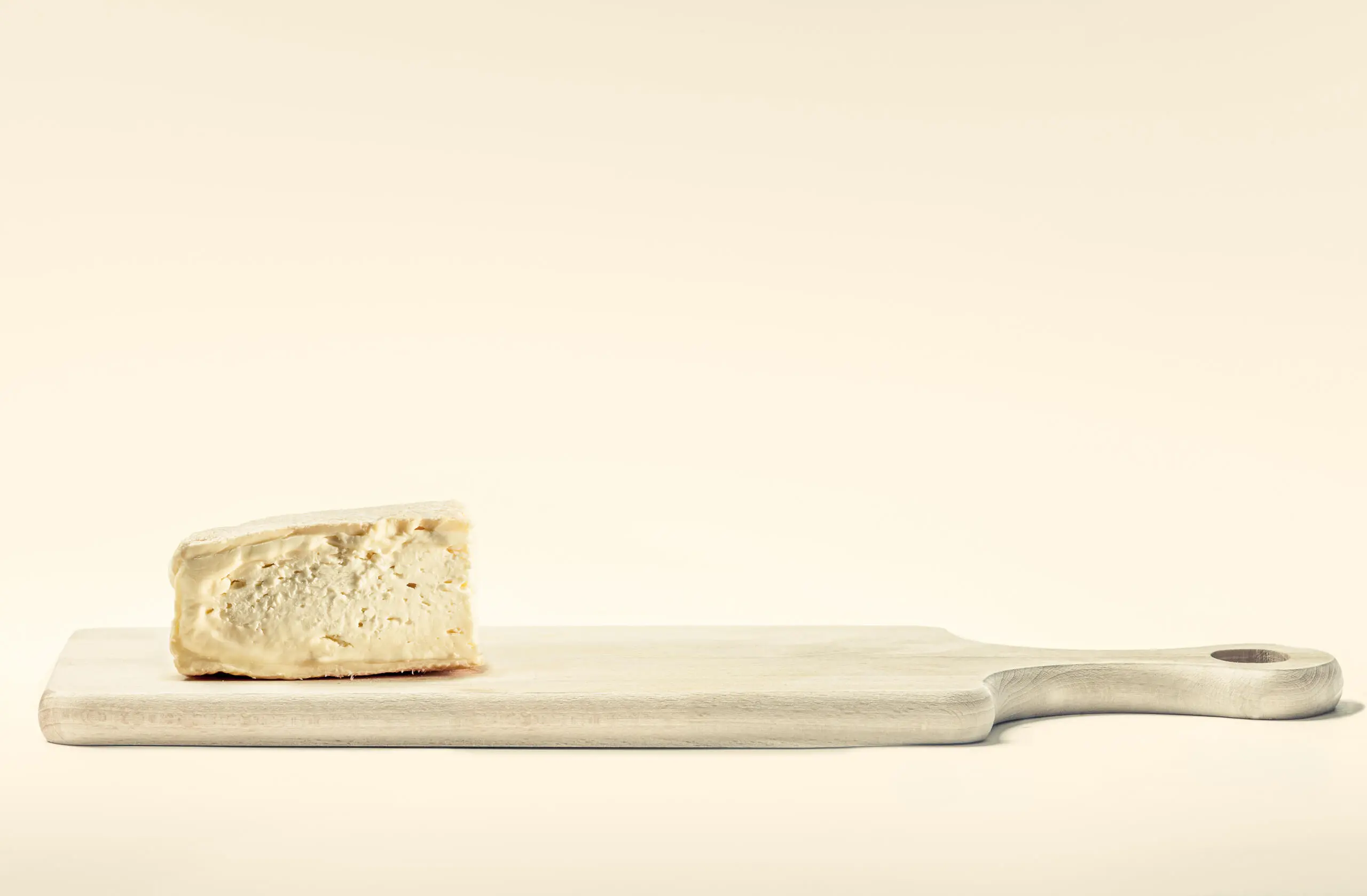 Ein Stück cremiger Weichkäse liegt auf einem hellen Holzschneidebrett vor einem neutralen, cremefarbenen Hintergrund.