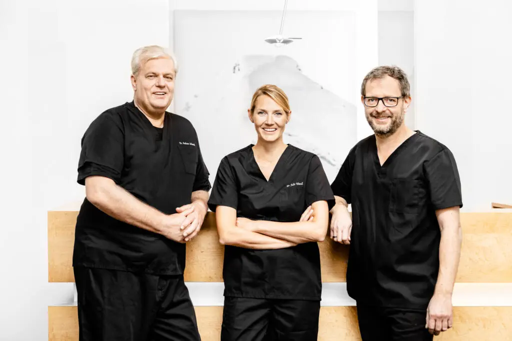 Drei medizinische Fachkräfte in schwarzer Arbeitskleidung stehen lächelnd hinter einem hellen Holzempfangstresen vor weißer Wand