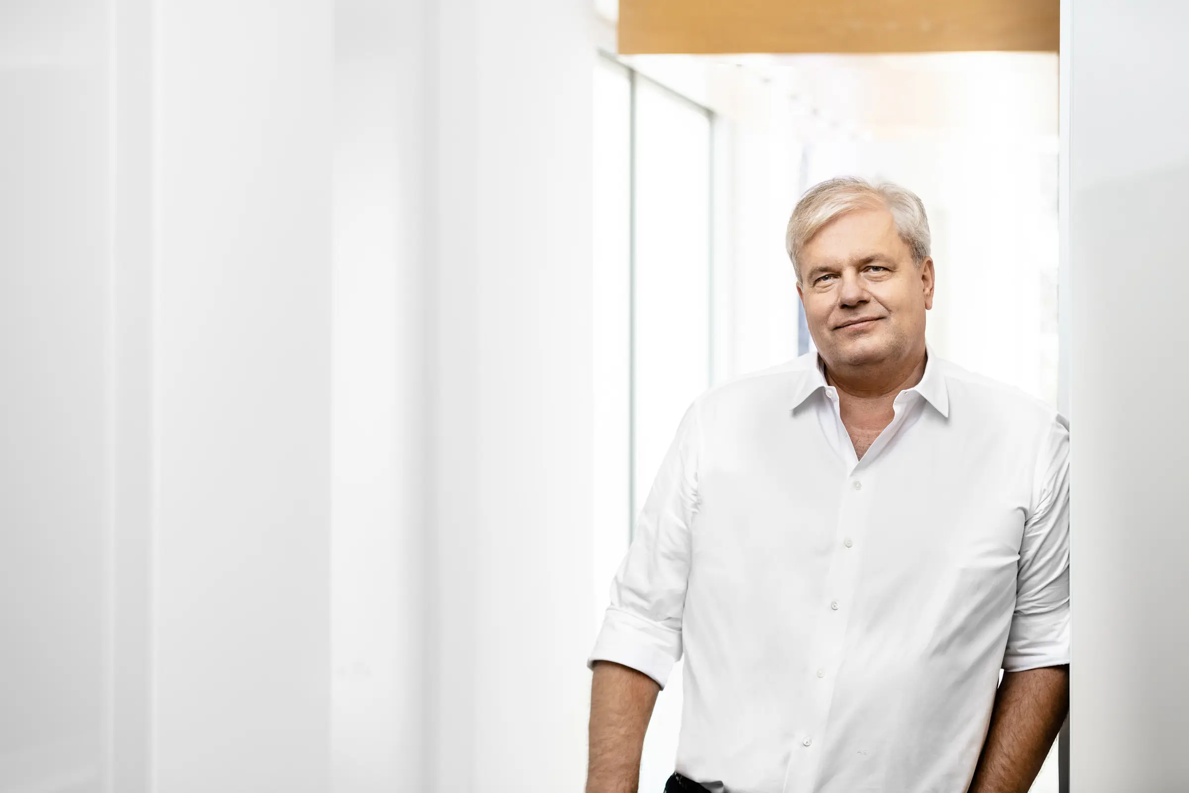 Porträt eines lächelnden Mannes mittleren Alters im weißen Hemd, der in einem hellen Praxisflur an einer Wand lehnt