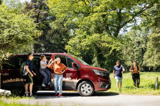 Group of people with backpacks exiting a red passenger van in a green outdoor setting for a team excursion